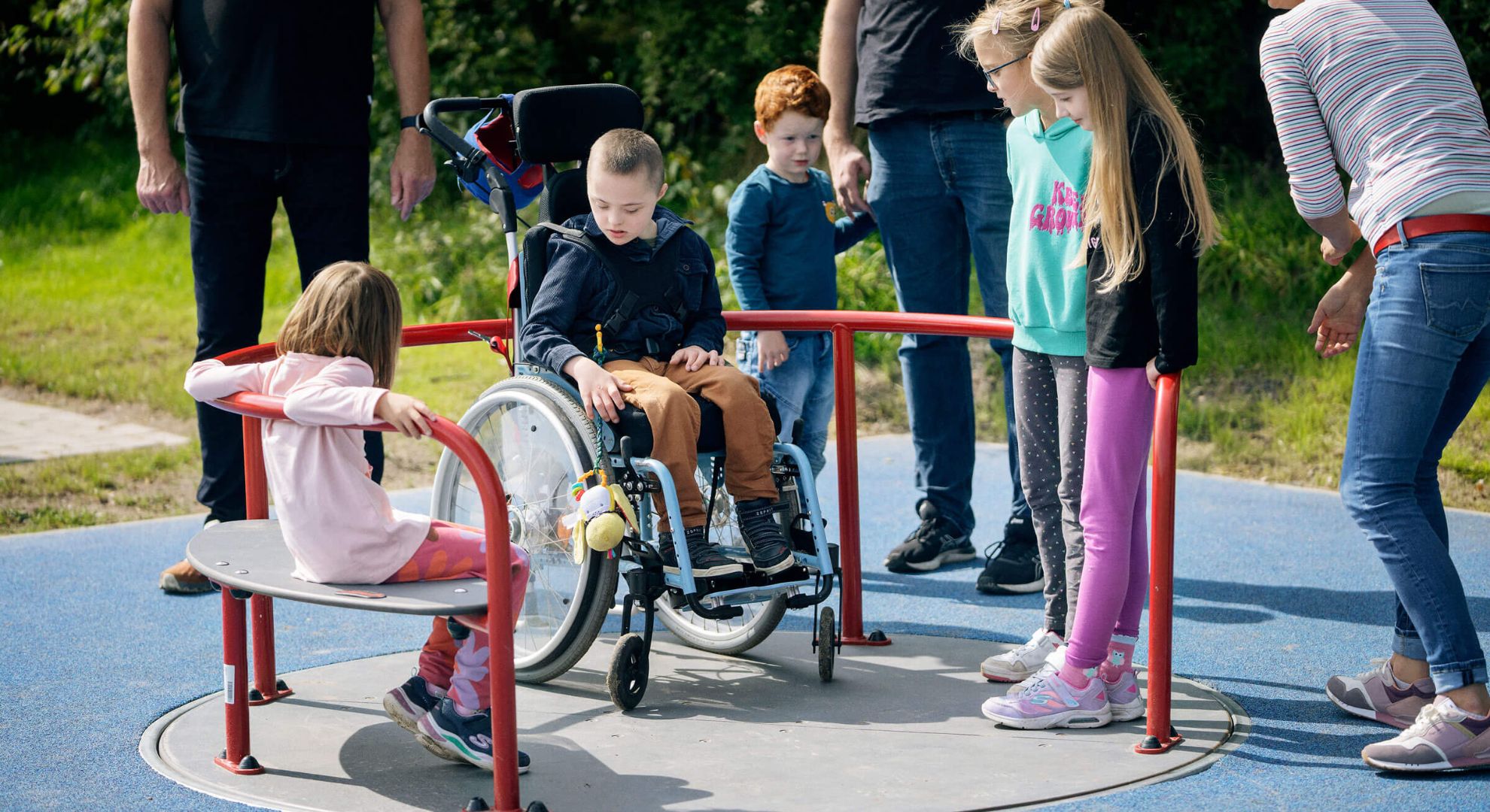 Child in wheelchair plays with diverse group at bright, inclusive playground. Green backdrop, blue accessible surface.